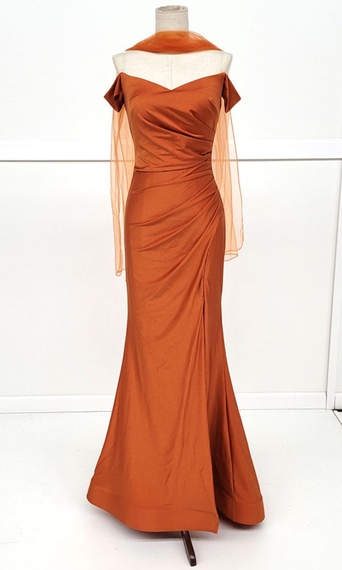 The Off-the-Shoulder Long Prom Dress: Chicas YG5025 in burnt orange features a ruched bodice, draped waist, and sheer shawl, shown on a dress form against a simple backdrop.